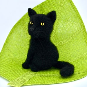 May include: A black knitted cat toy sitting on a green leaf-shaped mat. The cat has yellow eyes and a fluffy tail.