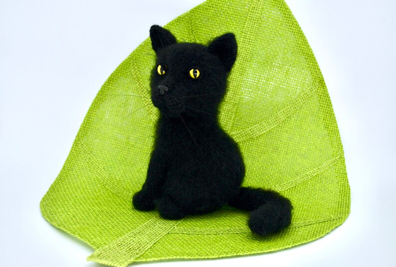 May include: A black knitted cat toy sitting on a green leaf-shaped mat. The cat has yellow eyes and a fluffy tail.