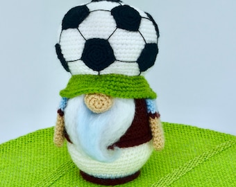 May include: A crocheted gnome wearing a white and black soccer ball hat, a green scarf, a brown vest, and a white shirt with blue trim. The gnome has a white beard and is standing on a green surface.