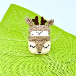 May include: A crocheted deer-themed AirPod case in tan, cream, and brown. The case features a deer face with closed eyes and a lid with antlers. The AirPods are visible inside the case. The case is on a green textured surface.