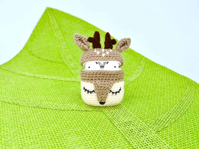 May include: A crocheted deer-themed AirPod case in tan, cream, and brown. The case features a deer face with closed eyes and a lid with antlers. The AirPods are visible inside the case. The case is on a green textured surface.