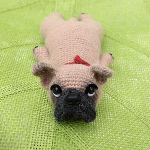 May include: A crocheted beige pug dog toy with black eyes, a black nose, and a red bow. The toy is lying on a green woven mat.