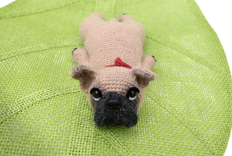 May include: A crocheted beige pug dog toy with black eyes, a black nose, and a red bow. The toy is lying on a green woven mat.
