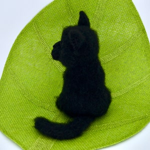 May include: A black needle felted cat sitting on a green leaf-shaped mat. The cat is facing to the left and has a long tail.
