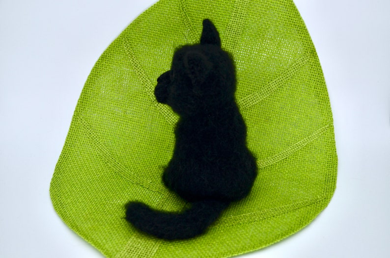 May include: A black needle felted cat sitting on a green leaf-shaped mat. The cat is facing to the left and has a long tail.