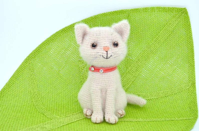 May include: A crocheted white cat with a red collar sitting on a green woven mat.