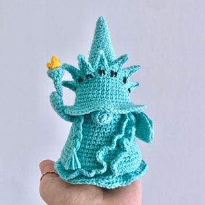 Puede incluir: Una figurita de crochet de la Estatua de la Libertad de color verde azulado. La figurita sostiene una antorcha en su mano derecha.