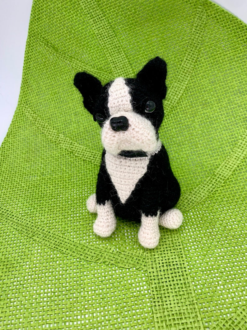 Pu&ograve; includere: Un giocattolo per cani Boston Terrier all'uncinetto con pelo nero e bianco. Il giocattolo &egrave; seduto su una superficie verde intrecciata.