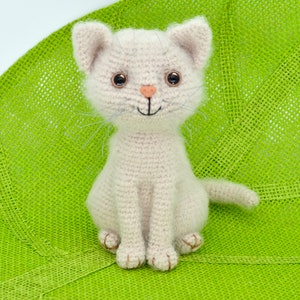 May include: A crocheted white cat with brown eyes and a pink nose sits on a green background. The cat is smiling and has a tail.