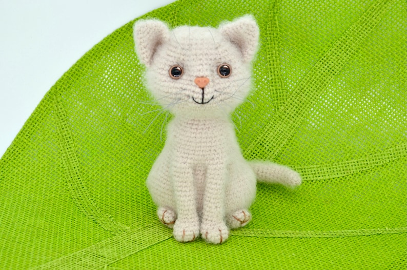 May include: A crocheted white cat with brown eyes and a pink nose sits on a green background. The cat is smiling and has a tail.