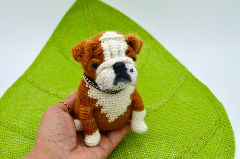 May include: A crocheted bulldog puppy toy with brown and white fur, wearing a black collar. The toy is sitting in a person's hand.