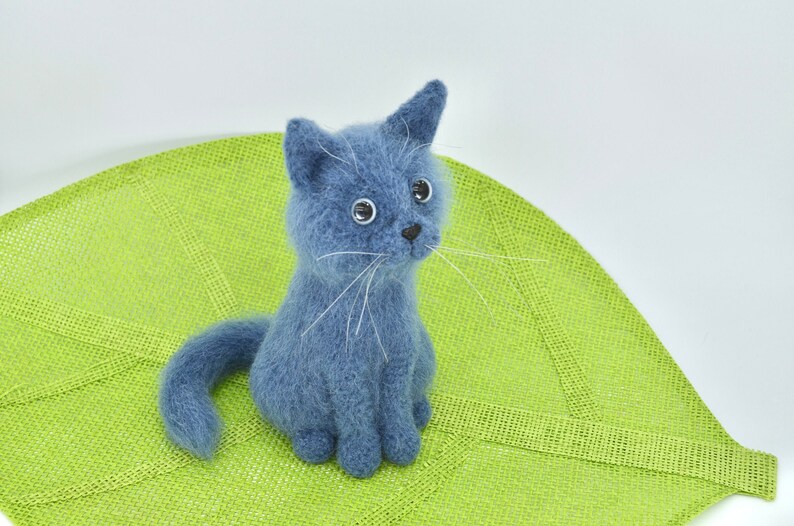 May include: A blue needle felted cat figurine sitting on a green crocheted surface.