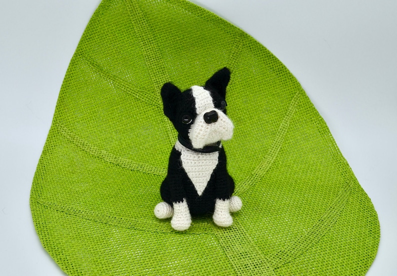Pu&ograve; includere: Un giocattolo da cane Boston Terrier in crochet nero e bianco seduto su un tappetino verde a forma di foglia.