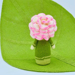 Op de afbeelding: Een gehaakte bloempop met een roze en witte bloemenkop en een groen lichaam. De pop zit op een groene, bladvormige mat.