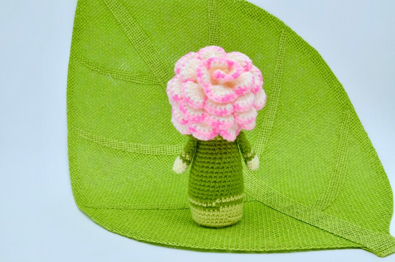 Op de afbeelding: Een gehaakte bloempop met een roze en witte bloemenkop en een groen lichaam. De pop zit op een groene, bladvormige mat.