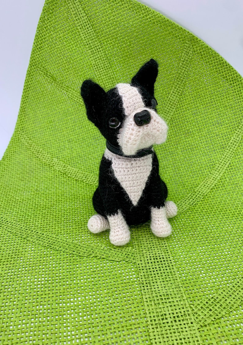 May include: A black and white crocheted Boston Terrier dog sitting on a green woven mat. The dog has black ears, a black nose, and white fur on its chest and paws.