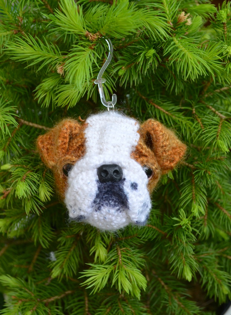 May include: A crocheted bulldog ornament hanging from a green pine tree branch. The bulldog is white with brown ears and a black nose.