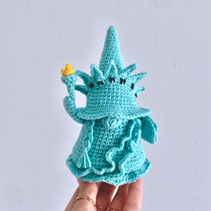 Puede incluir: Una figurita de crochet de la Estatua de la Libertad de color azul verdoso. La figurita sostiene una antorcha en su mano derecha y tiene una corona en la cabeza.