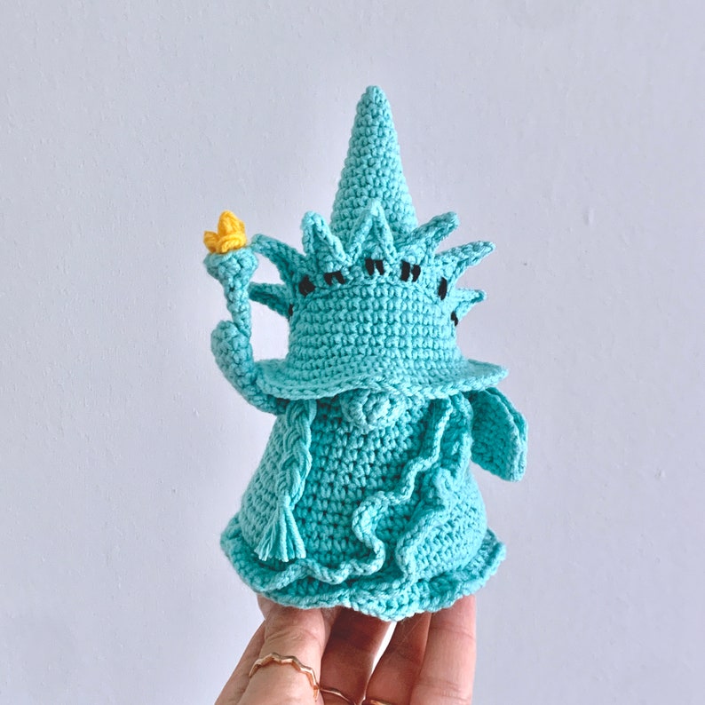 Puede incluir: Una figurita de crochet de la Estatua de la Libertad de color azul verdoso. La figurita sostiene una antorcha en su mano derecha y tiene una corona en la cabeza.