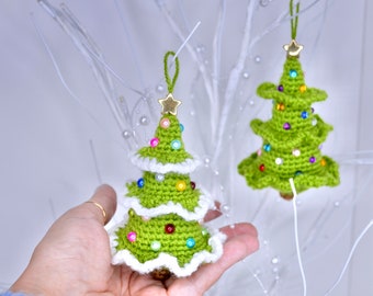 May include: Two green crocheted Christmas tree ornaments with white trim and colorful beads. The ornaments are hanging from a white branch.