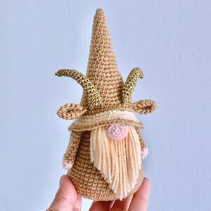 May include: A crocheted gnome with a brown hat, a pink nose, and a long, cream-coloured beard. The gnome has gold glitter horns.