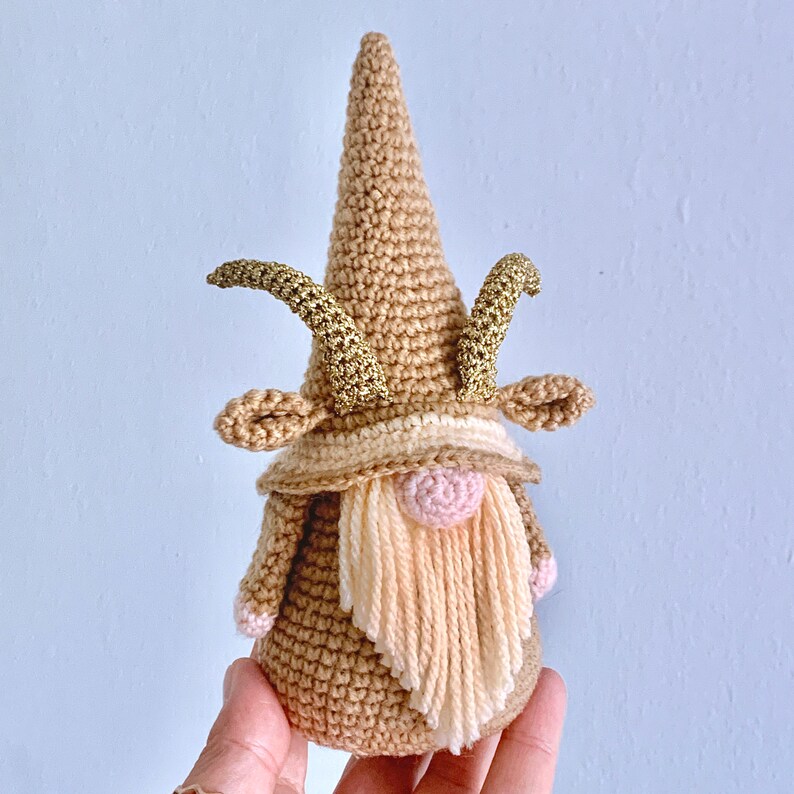 May include: A crocheted gnome with a brown hat, a pink nose, and a long, cream-coloured beard. The gnome has gold glitter horns.