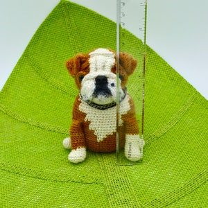 May include: A crocheted brown and white bulldog puppy toy sitting on a green burlap surface. The toy is approximately 6 inches tall.