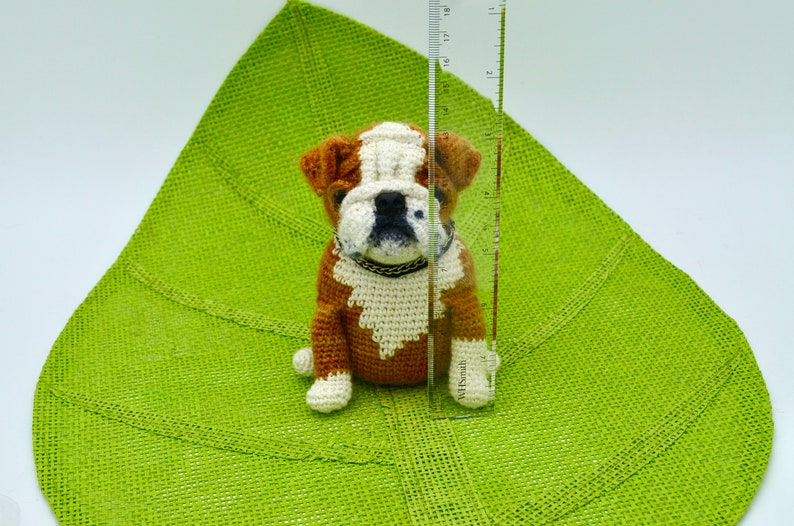 May include: A crocheted brown and white bulldog puppy toy sitting on a green burlap surface. The toy is approximately 6 inches tall.