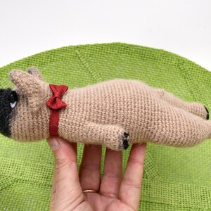 May include: A crocheted beige dog toy with black eyes and a red bow tie. The toy is lying on its back with its paws in the air.