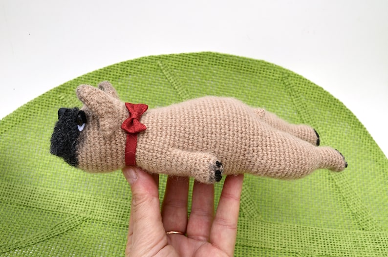 May include: A crocheted beige dog toy with black eyes and a red bow tie. The toy is lying on its back with its paws in the air.