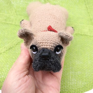 May include: A crocheted beige French bulldog plush toy with black eyes and a black nose. The toy is being held in a hand.