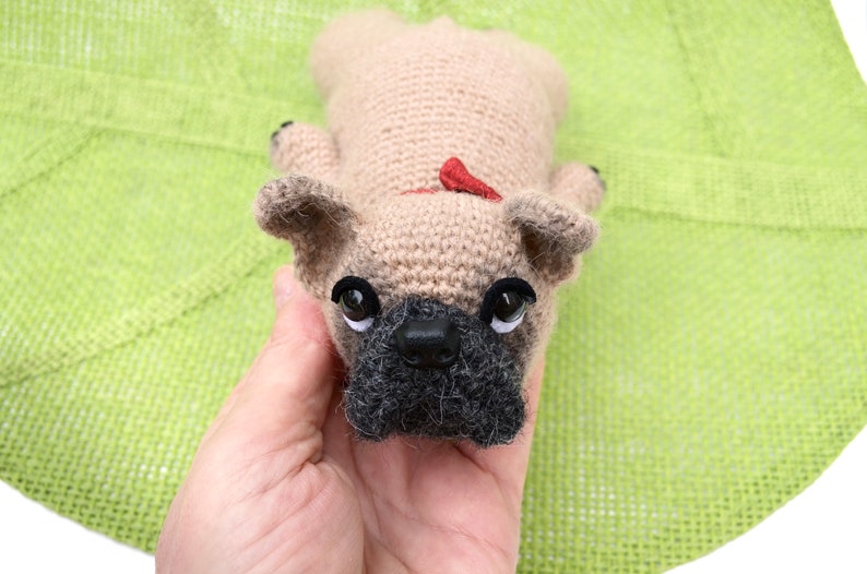 May include: A crocheted beige French bulldog plush toy with black eyes and a black nose. The toy is being held in a hand.