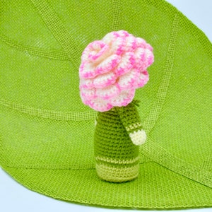Op de afbeelding: Een gehaakte bloempot met een roze en witte bloem erop. De pot is groen en heeft een witte streep rond het midden.