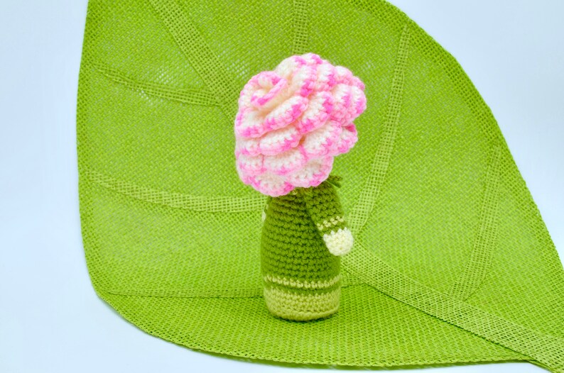 Op de afbeelding: Een gehaakte bloempot met een roze en witte bloem erop. De pot is groen en heeft een witte streep rond het midden.
