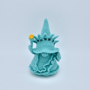 Puede incluir: Un gnomo de crochet con un sombrero azul con forma de la Estatua de la Libertad. El gnomo sostiene una flor amarilla en su mano.