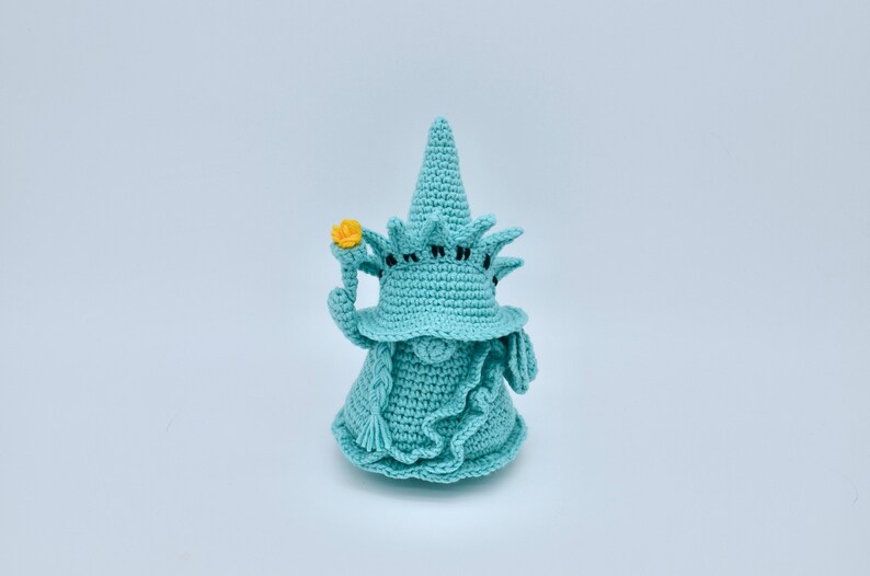 Puede incluir: Un gnomo de crochet con un sombrero azul con forma de la Estatua de la Libertad. El gnomo sostiene una flor amarilla en su mano.