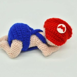 May include: Crocheted baby doll wearing a blue diaper and a red hat with a white emblem. The doll is lying on its stomach, showcasing the handmade details and vibrant colors. A unique, handcrafted item.