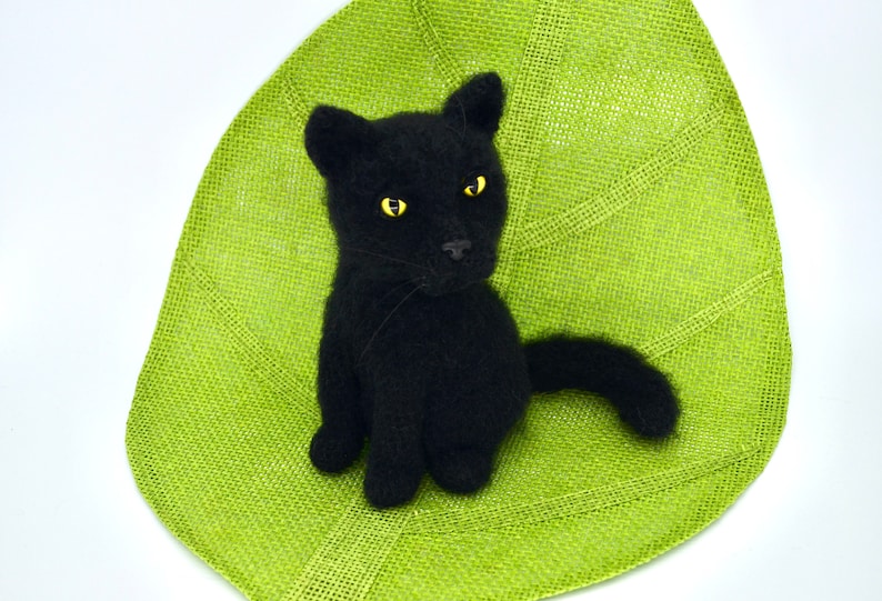 May include: A black knitted cat with yellow eyes sitting on a green burlap leaf.