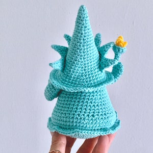 Puede incluir: Un gnomo de crochet azul turquesa con un sombrero puntiagudo y una flor amarilla en la parte superior. El gnomo est&aacute; de pie sobre una mano.