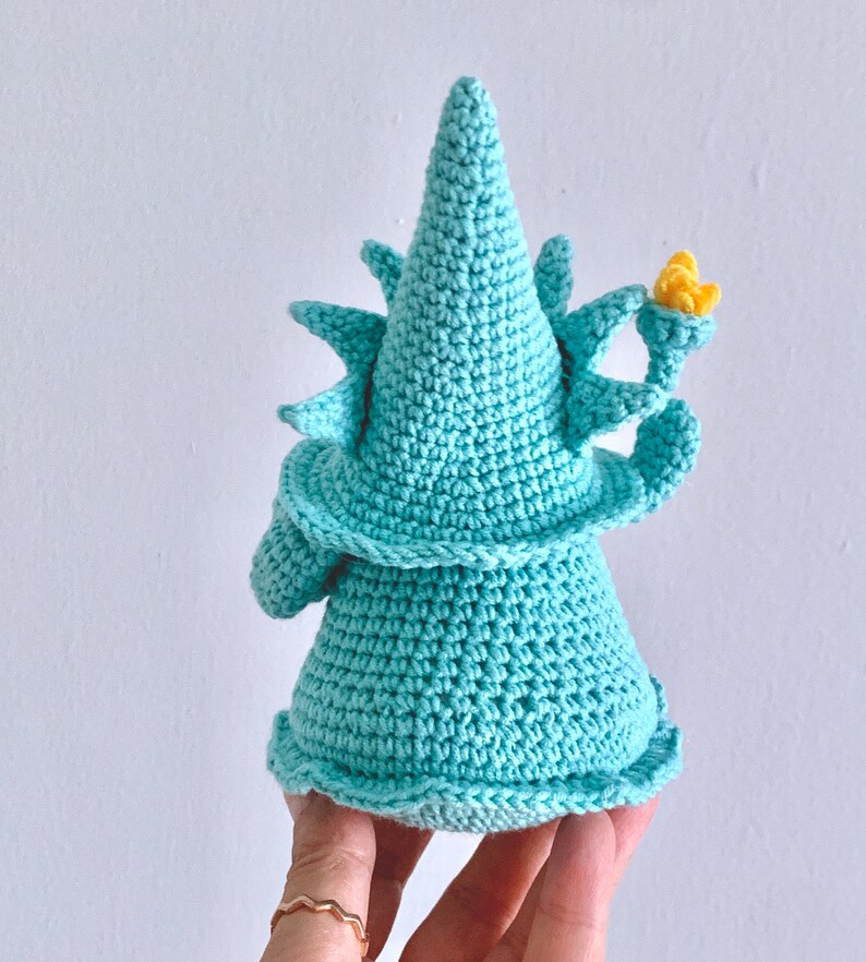 Puede incluir: Un gnomo de crochet azul turquesa con un sombrero puntiagudo y una flor amarilla en la parte superior. El gnomo est&aacute; de pie sobre una mano.