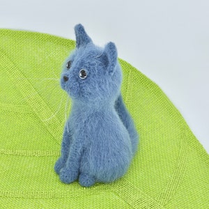 May include: A blue knitted cat toy sitting on a green leaf-shaped mat. The cat has white whiskers and black eyes.