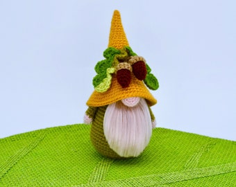 May include: A handmade crochet gnome doll with a yellow hat, green body, and a long white beard. The hat is adorned with green leaves and two brown acorns. The gnome is sitting on a green textured surface.