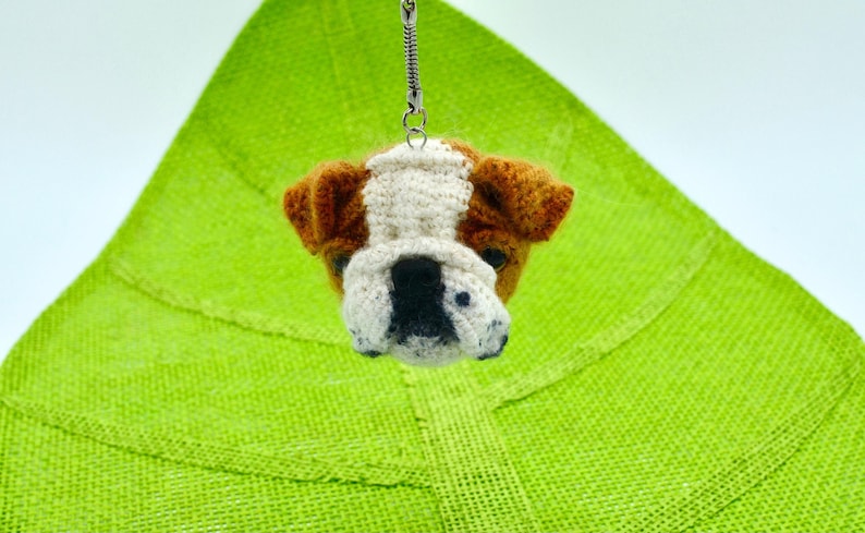 May include: A crocheted bulldog keychain with brown, white, and black fur. The keychain is hanging from a silver chain.