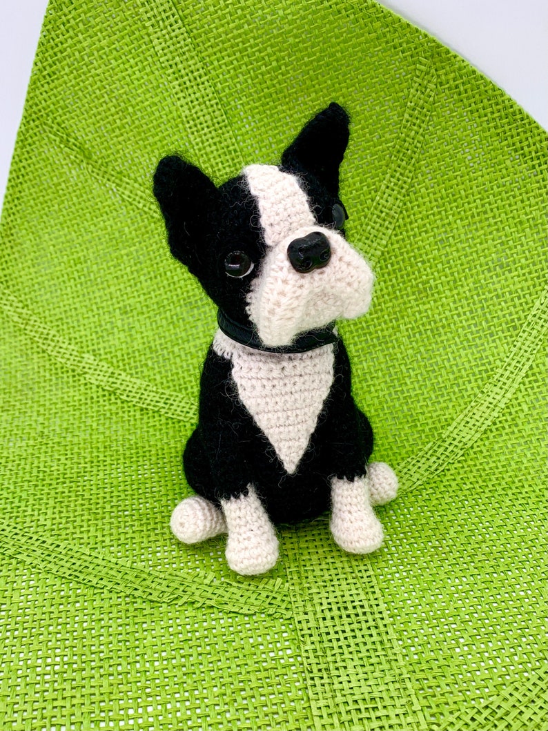 Pu&ograve; includere: Un giocattolo per cani Boston Terrier all'uncinetto, nero e bianco con un collare nero, seduto su una superficie verde intrecciata.