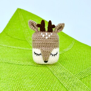 May include: A crocheted deer-shaped container with a brown and white color scheme. The deer has closed eyes, small antlers, and white spots. The container is set against a bright green textured background.