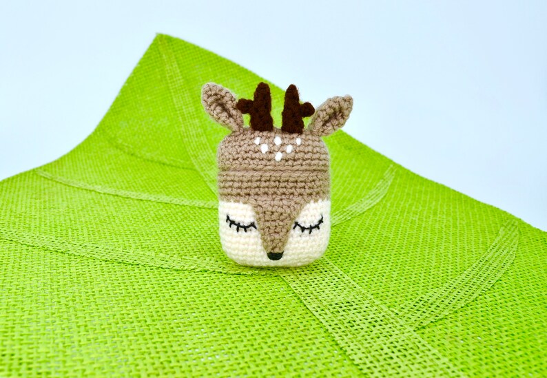 May include: A crocheted deer-shaped container with a brown and white color scheme. The deer has closed eyes, small antlers, and white spots. The container is set against a bright green textured background.