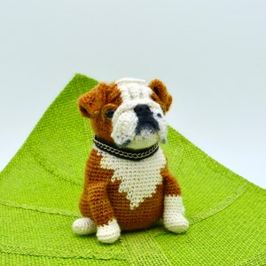 May include: A crocheted bulldog plush toy with brown and white fur, wearing a black collar with a silver chain, sitting on a green fabric surface.