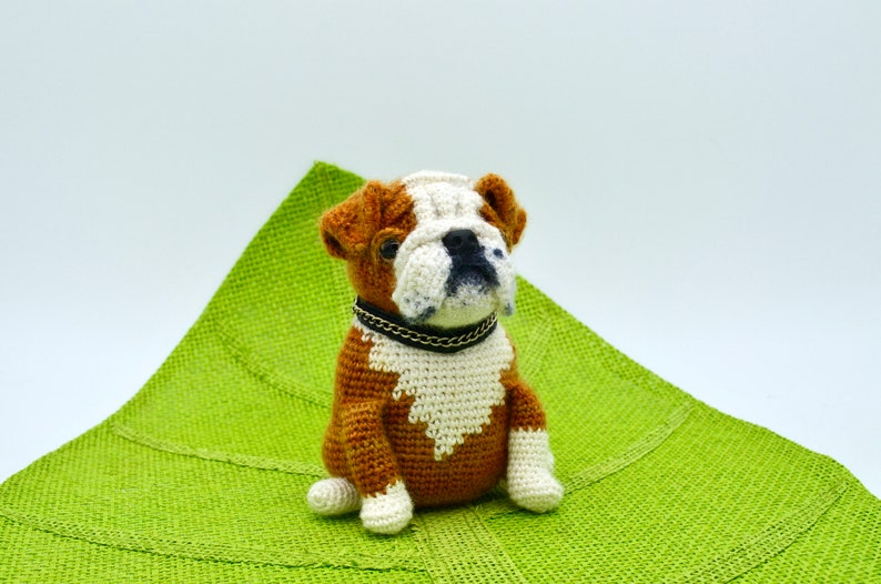 May include: A crocheted bulldog plush toy with brown and white fur, wearing a black collar with a silver chain, sitting on a green fabric surface.