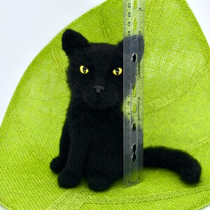 May include: A black needle felted cat with yellow eyes sitting on a green background. A ruler is next to the cat for size reference.