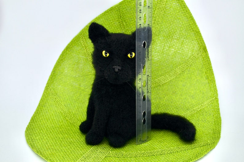 May include: A black needle felted cat with yellow eyes sitting on a green background. A ruler is next to the cat for size reference.